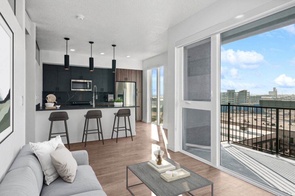 Modern apartment interior with a gray sofa, kitchen with bar stools, and a balcony overlooking city buildings under a blue sky with clouds.