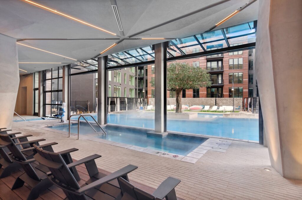 Indoor pool area with lounge chairs overlooks an outdoor swimming pool through large glass windows; a tree and modern apartment buildings are visible outside.