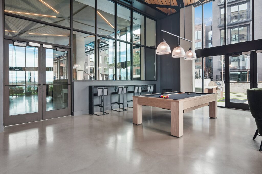 Modern recreation room with a pool table, bar seating, large windows, and pendant lights, featuring polished concrete floors and an outdoor view.