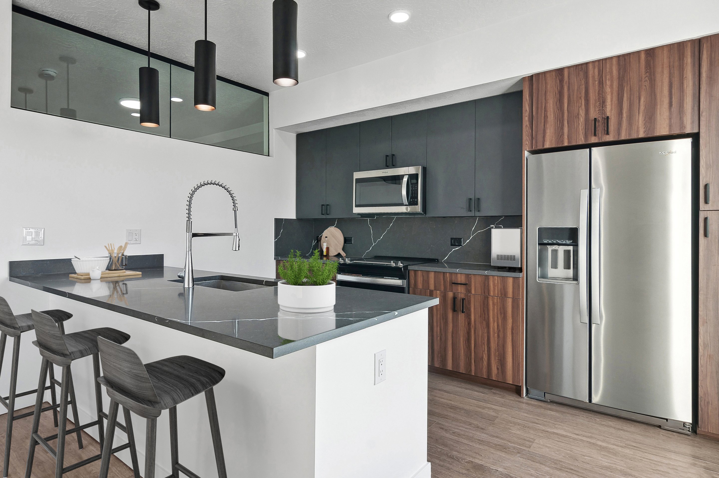 Modern kitchen in Home - Lake Cameron with black countertops, wood cabinetry, stainless steel appliances, an island with bar stools, and a potted plant on the counter.