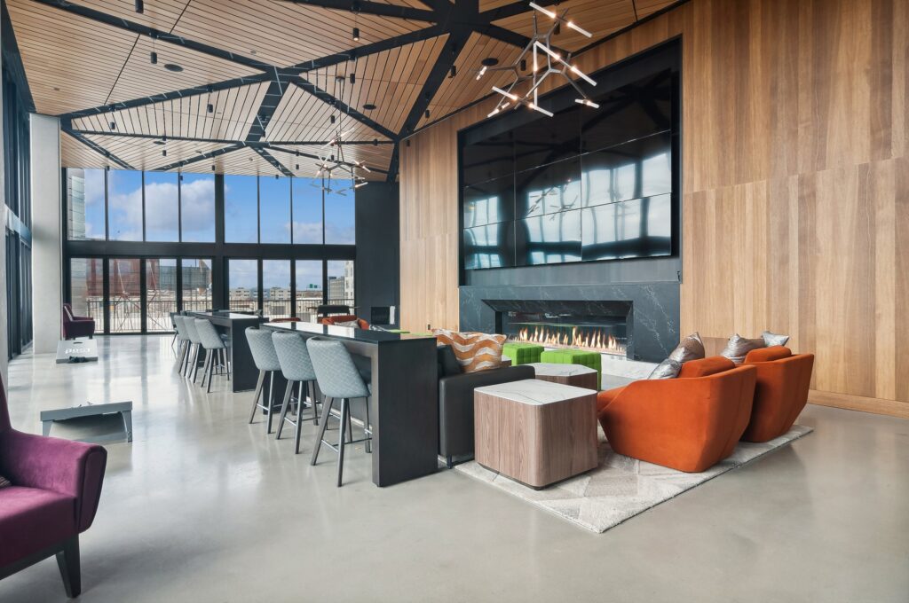 Modern lounge area with a long bar, orange chairs, a fireplace, large screen, wood-paneled walls, and floor-to-ceiling windows overlooking the city.