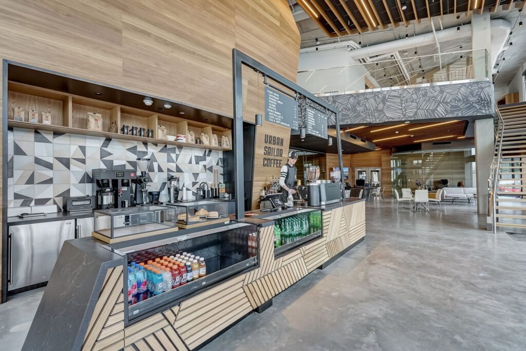 Modern coffee shop interior with a geometric counter, beverage display, coffee machines, and a seating area in a spacious, open setting with high ceilings and wooden accents.