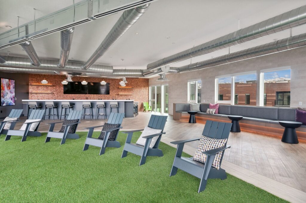 Modern indoor lounge with artificial grass, black chairs with cushions, a long bar counter, and large windows letting in natural light.
