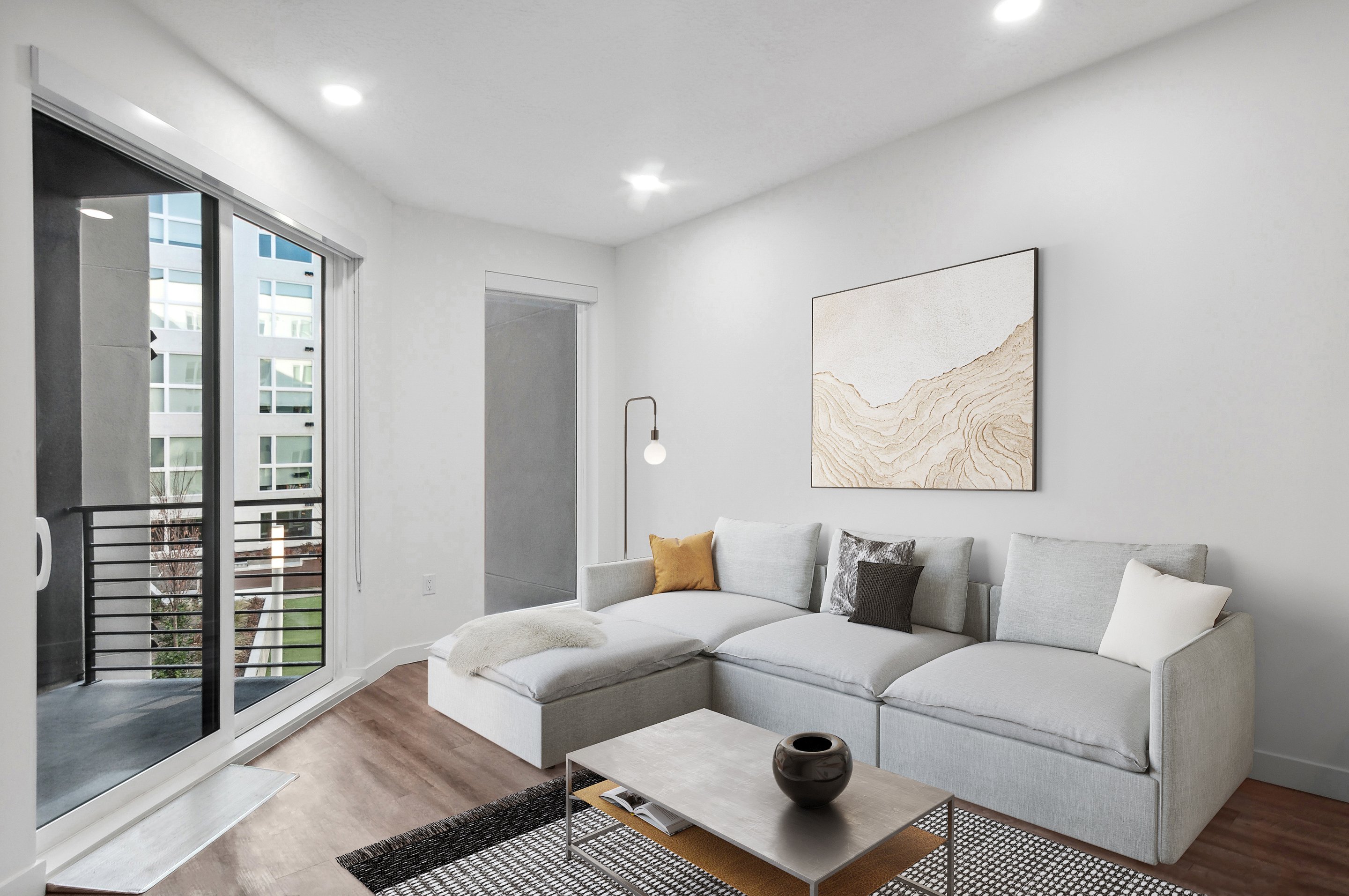 Modern living room at Home - Lake Cameron featuring a large gray sectional sofa, abstract wall art, floor lamp, coffee table, and balcony access through sliding glass doors.