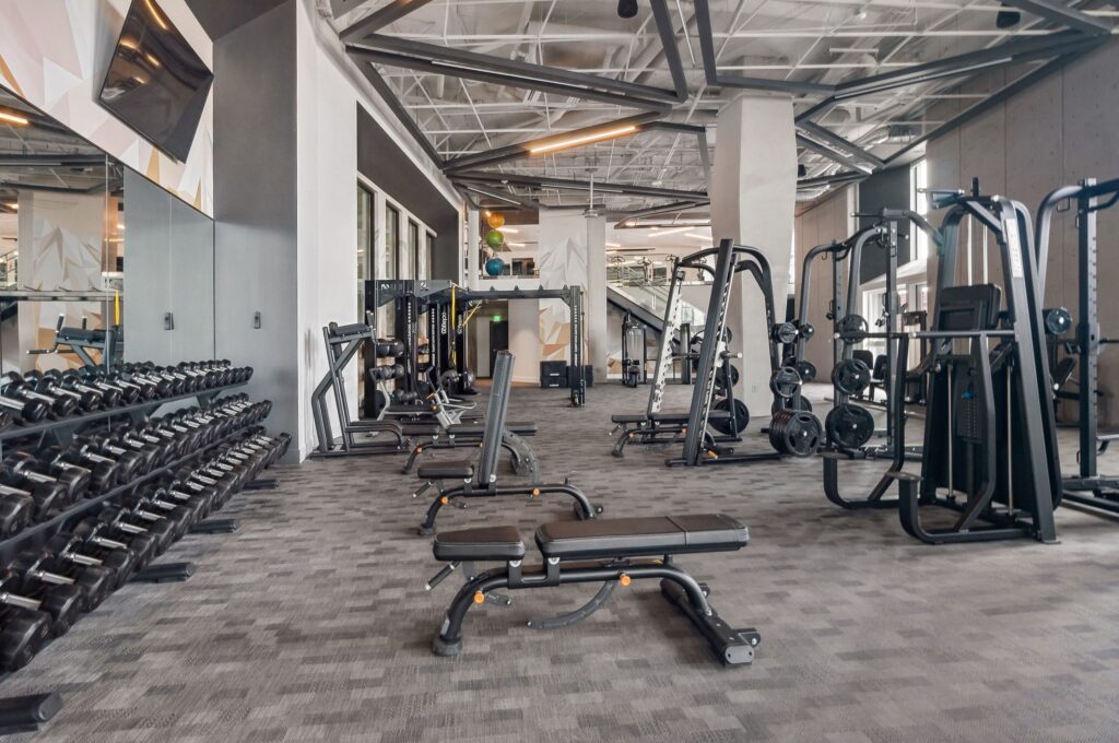 Modern gym with weight benches, dumbbells, and various strength training equipment on a gray carpeted floor. The space is well-lit with high ceilings and large mirrors.