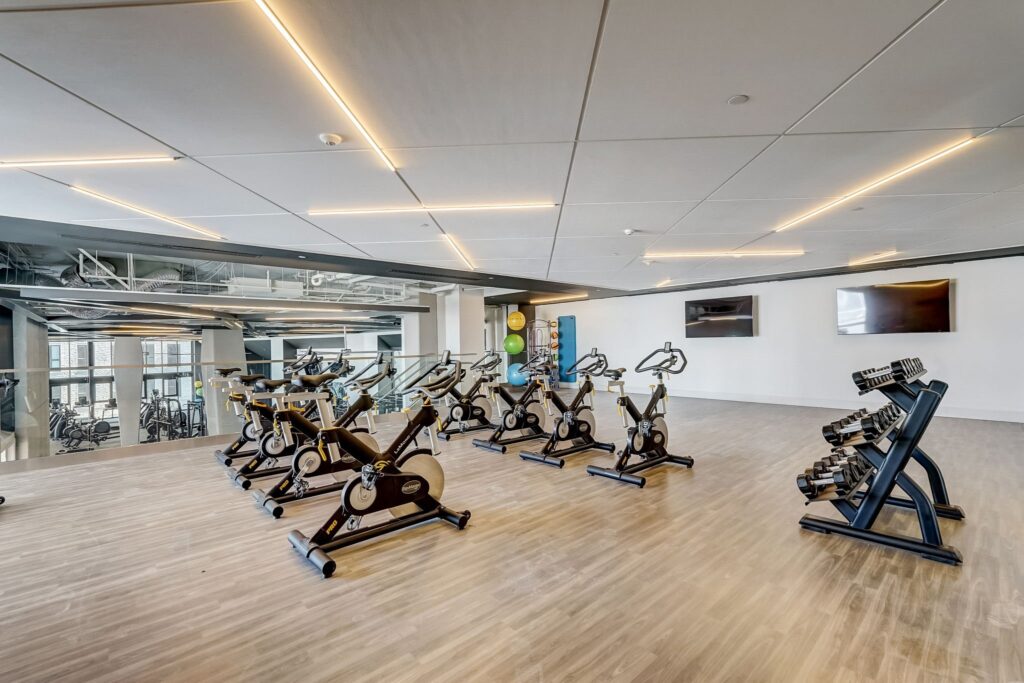 A modern fitness studio with several stationary bikes, free weights on racks, wood flooring, and wall-mounted TVs.