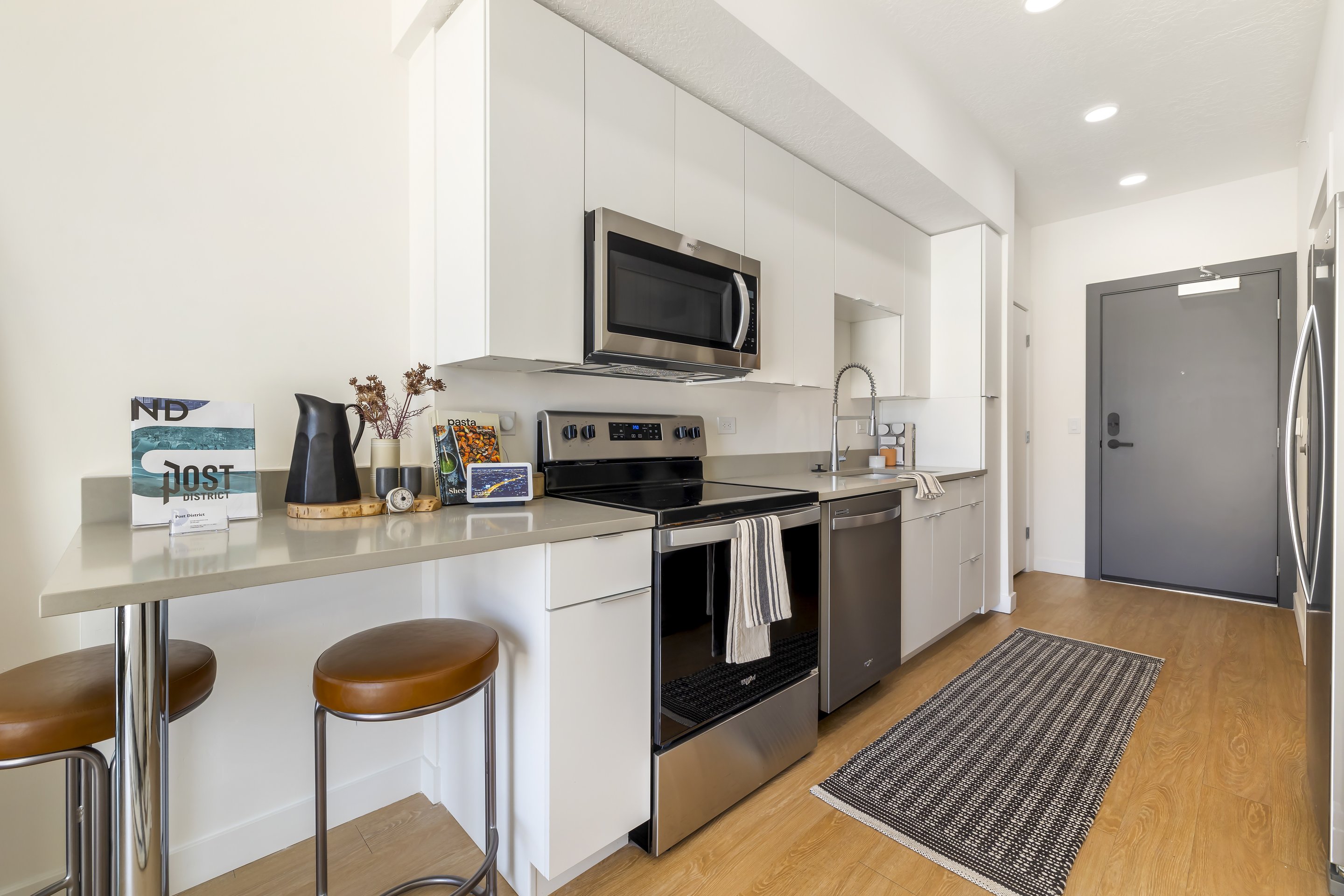 Modern apartment kitchen in Home - Lake Cameron with white cabinets, stainless steel appliances, a small peninsula with two stools, and a gray rug on wood flooring.