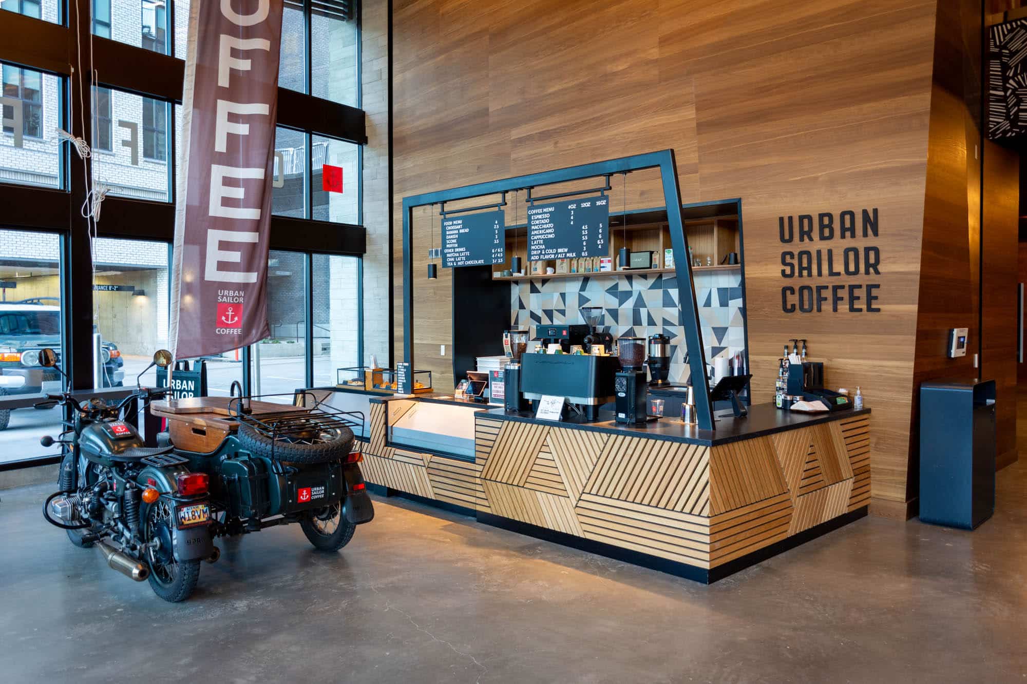 A modern coffee shop named Urban Sailor Coffee features a geometric wood counter, overhead menu, and vintage motorcycle parked indoors near large windows, creating an inviting vibe reminiscent of Home - Lake Cameron.