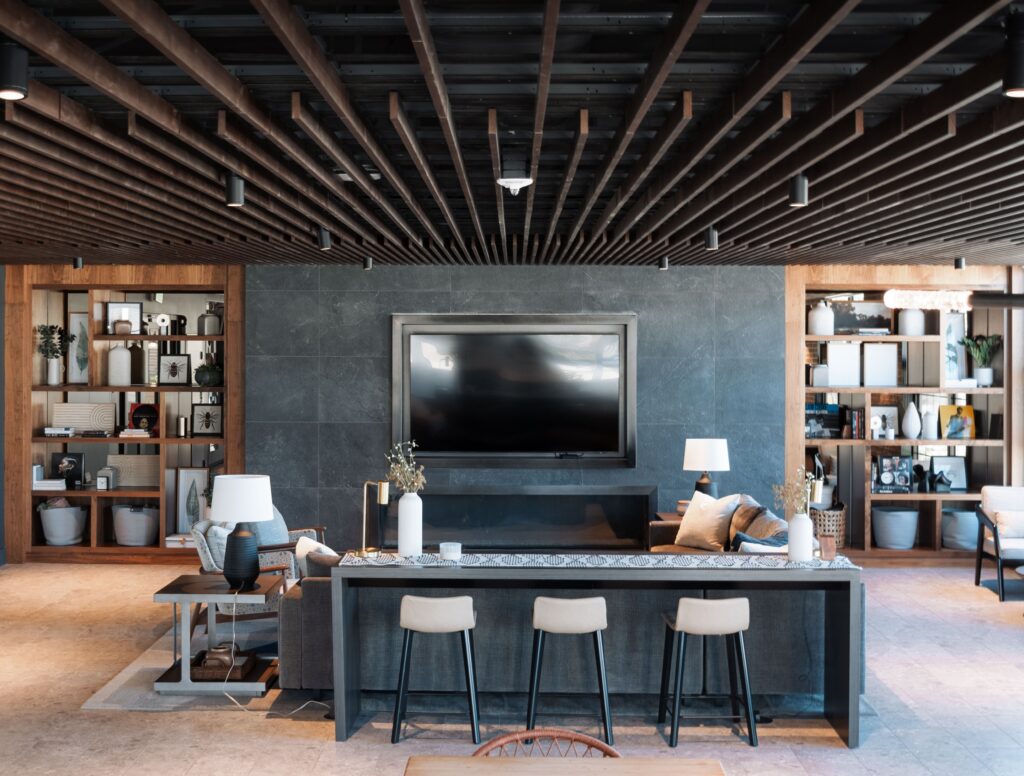 Modern living room with a large TV on a dark feature wall, a long table with bar stools, couches, and open shelves filled with decorative items and plants.