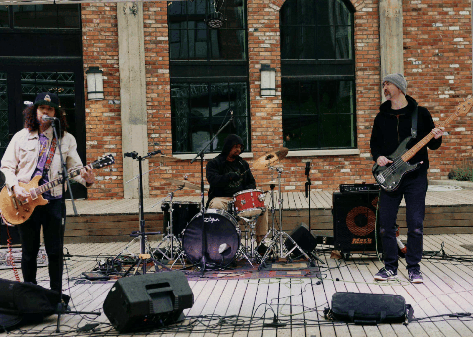 Three musicians perform on an outdoor wooden stage with drums, electric guitar, and bass guitar in front of the brick Home - Post District Residences building with large windows.