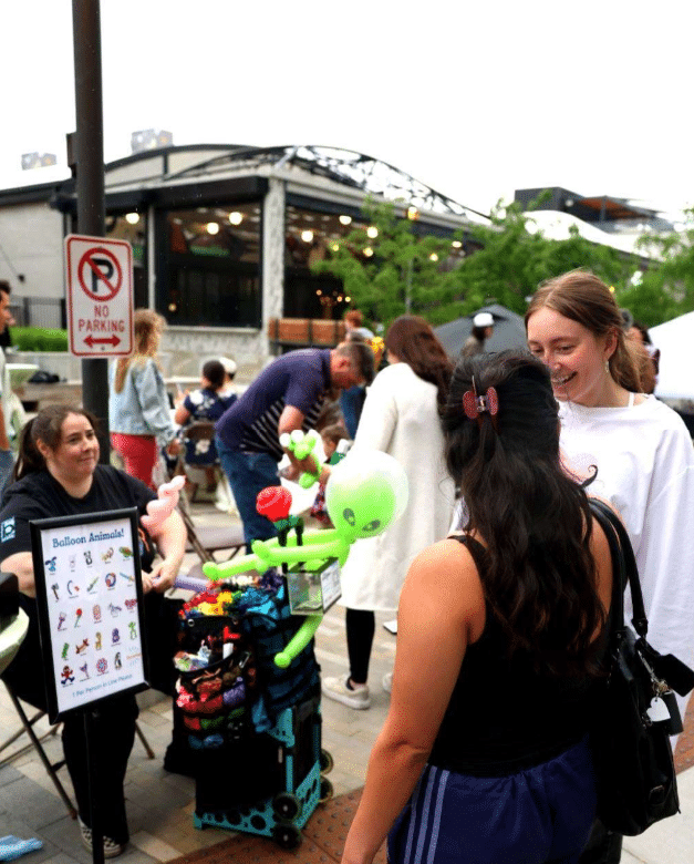 People gather at an outdoor event near a balloon artist's stand with a display of balloon animal options by Home - Post District Residences. A "No Parking" sign is visible next to the crowd.